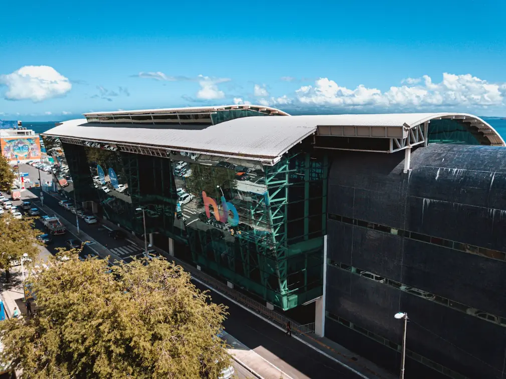 Vista aérea do prédio do Hub Salvador, com cobertura curva, estrutura metálica verde e o letreiro do hub na fachada envidraçada, com o mar ao fundo