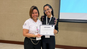 Duas mulheres sorridentes, lado a lado, seguram juntas um certificado com o título 'Prêmio ABEPRO'. A mulher à esquerda usa uma blusa branca e crachá, e a mulher à direita usa óculos, blusa preta bordada com flores e também crachá. Ao fundo, uma parede bege e uma tela de projeção azul.
