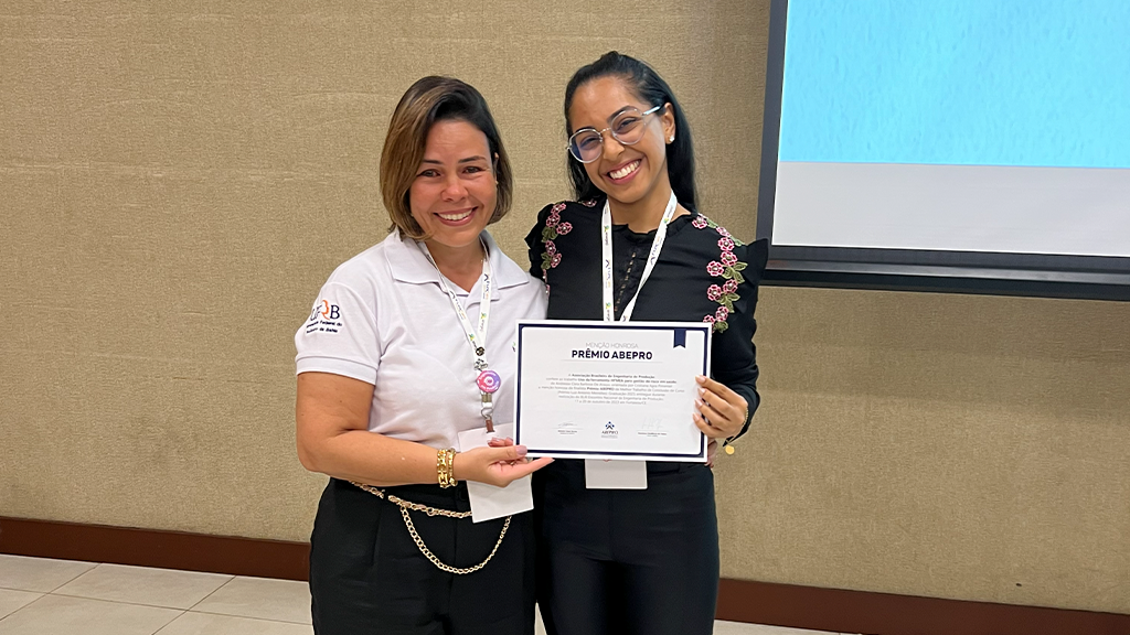 Duas mulheres sorridentes, lado a lado, seguram juntas um certificado com o título 'Prêmio ABEPRO'. A mulher à esquerda usa uma blusa branca e crachá, e a mulher à direita usa óculos, blusa preta bordada com flores e também crachá. Ao fundo, uma parede bege e uma tela de projeção azul.